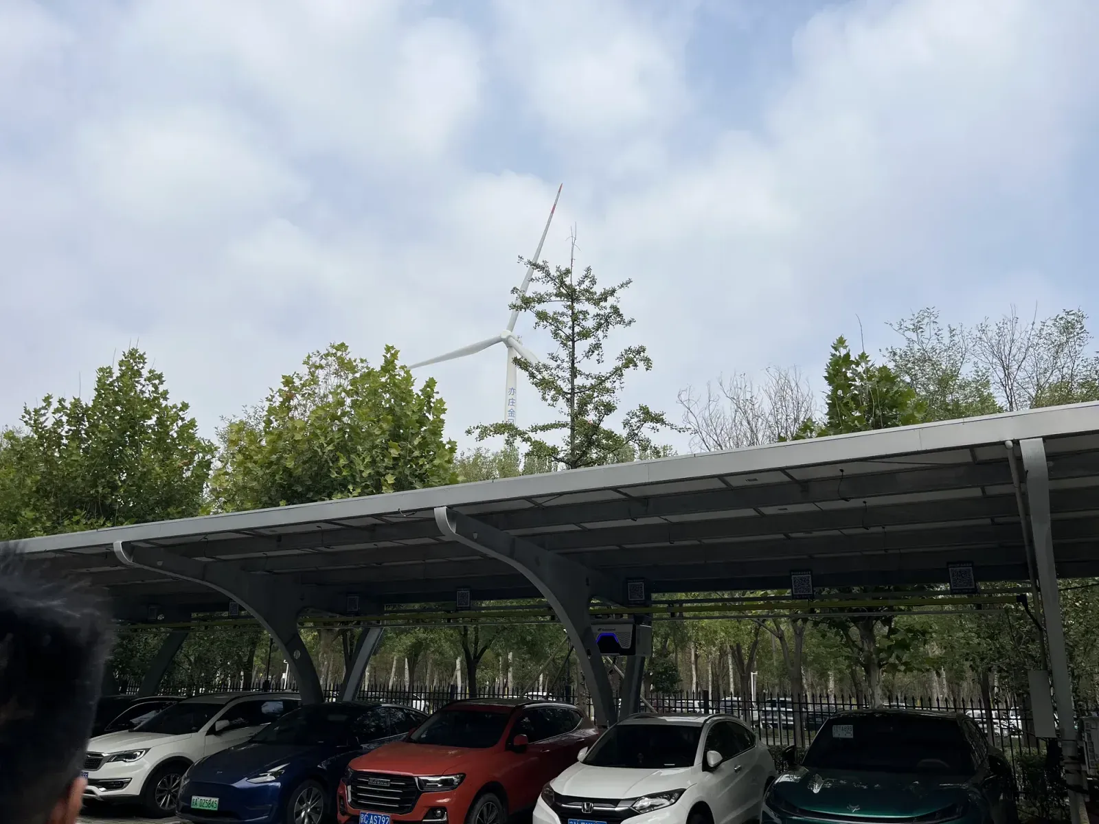 Electric cars charging beneath solar panels alongside a wind turbine